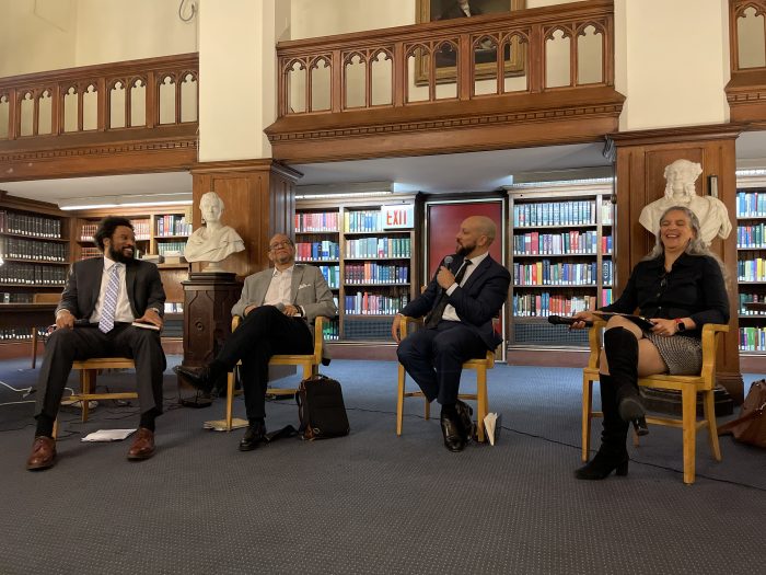 three men -- Nkosi Anderson a young Black man, Obery Hendricks an older Black man, and Josef Sorett an adult Black man, and Andrea White a Black woman with long grey hair, sit side by side in wooden chairs in a library room lined with books