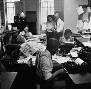 African American women and men in a newsroom with typewriters and newspapers on desks